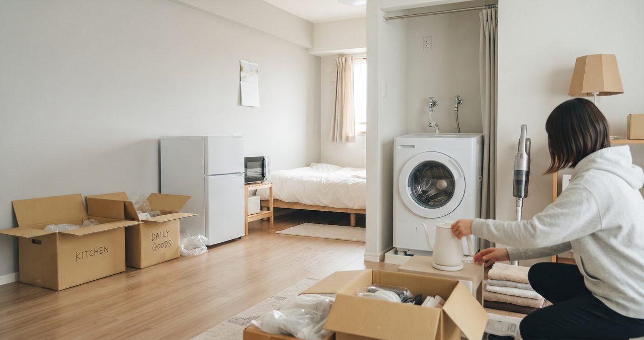 引っ越し直後の部屋で、段ボールを開けて荷物を出す女性と、冷蔵庫・洗濯機・掃除機