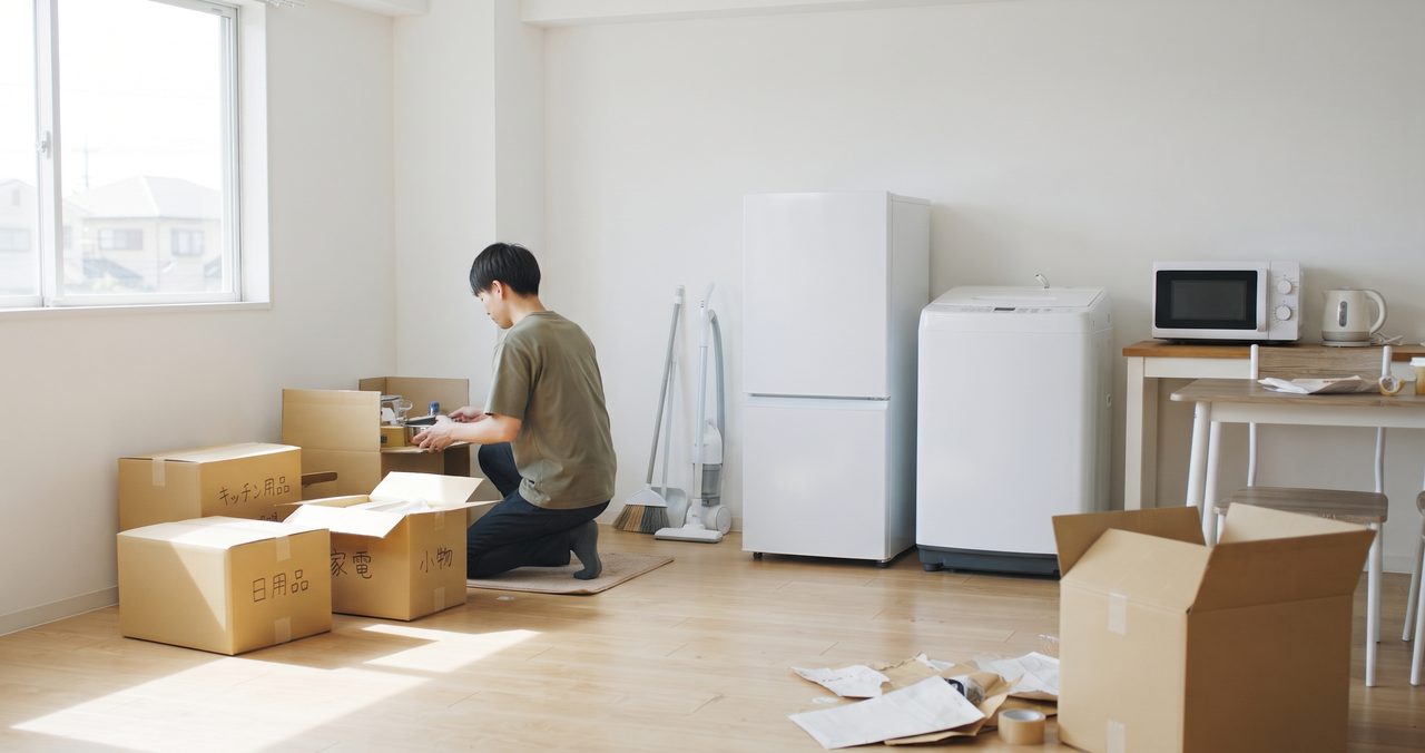 白い冷蔵庫と洗濯機が置かれた部屋で、段ボールを開けて荷ほどきをする男性