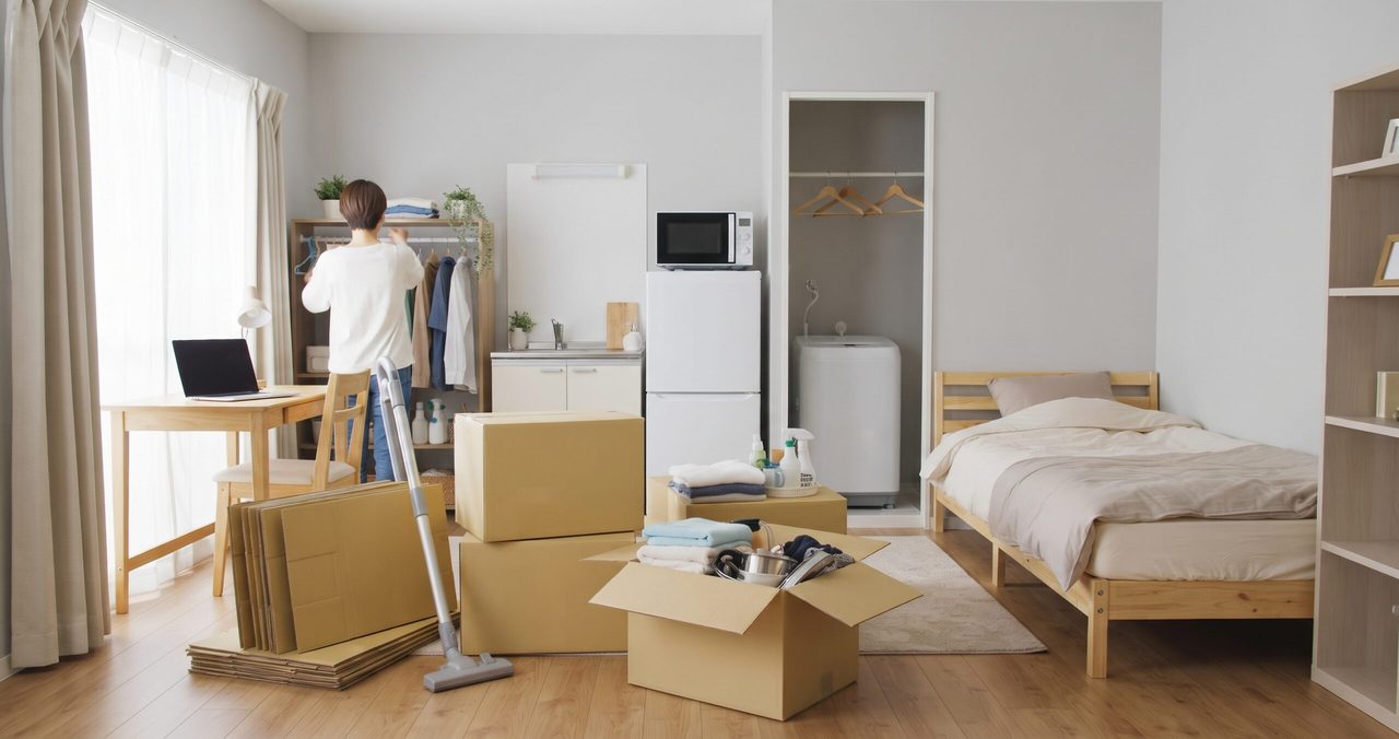 ベッド、冷蔵庫、洗濯機、机のある部屋で、掃除機をかけながら荷物を片付ける男性