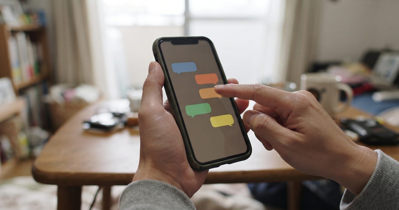 単身赴任先の部屋でスマートフォンを片手に持ち、メッセージアプリの画面を指でタップして家族に連絡している手元の様子